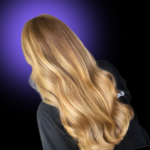 Long, multi-dimensional honey blonde and golden blonde balayage hair color. Seamless color melt styled into smooth luxury waves, shot from behind against a black background with a purple vignette.