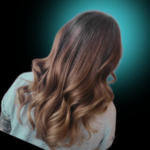 Brunette to caramel blonde ombré hair color transition. Long hair styled with voluminous, bouncy curls at the ends, displayed against a dark background with a teal light glow.